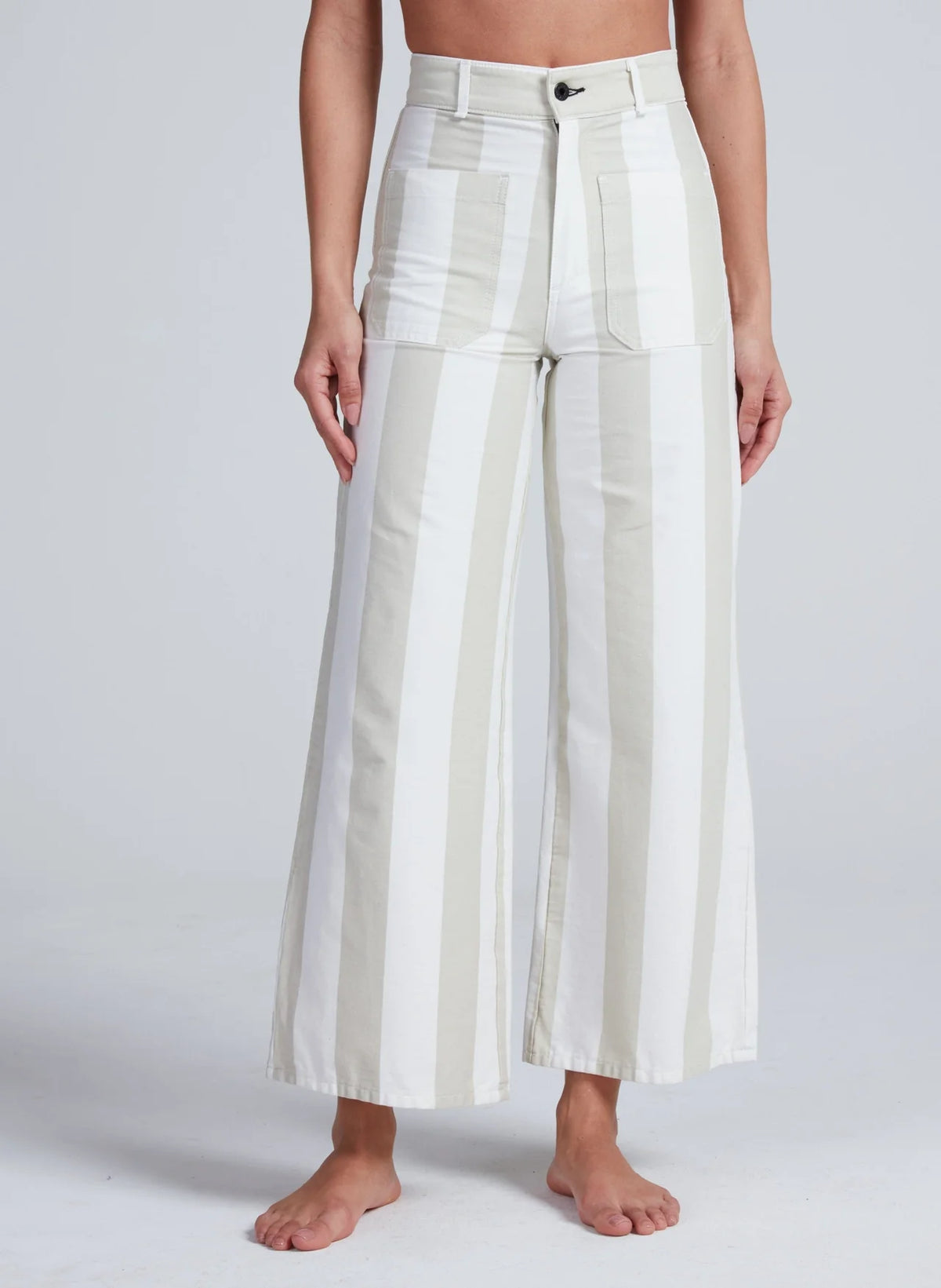 A person stands barefoot in ASKK’s Cropped Sailor pants, featuring high rise, wide legs, large front pockets, and bold white and light beige vertical stripes. The stretch twill adds comfort. Their torso appears partially against a plain light background.
