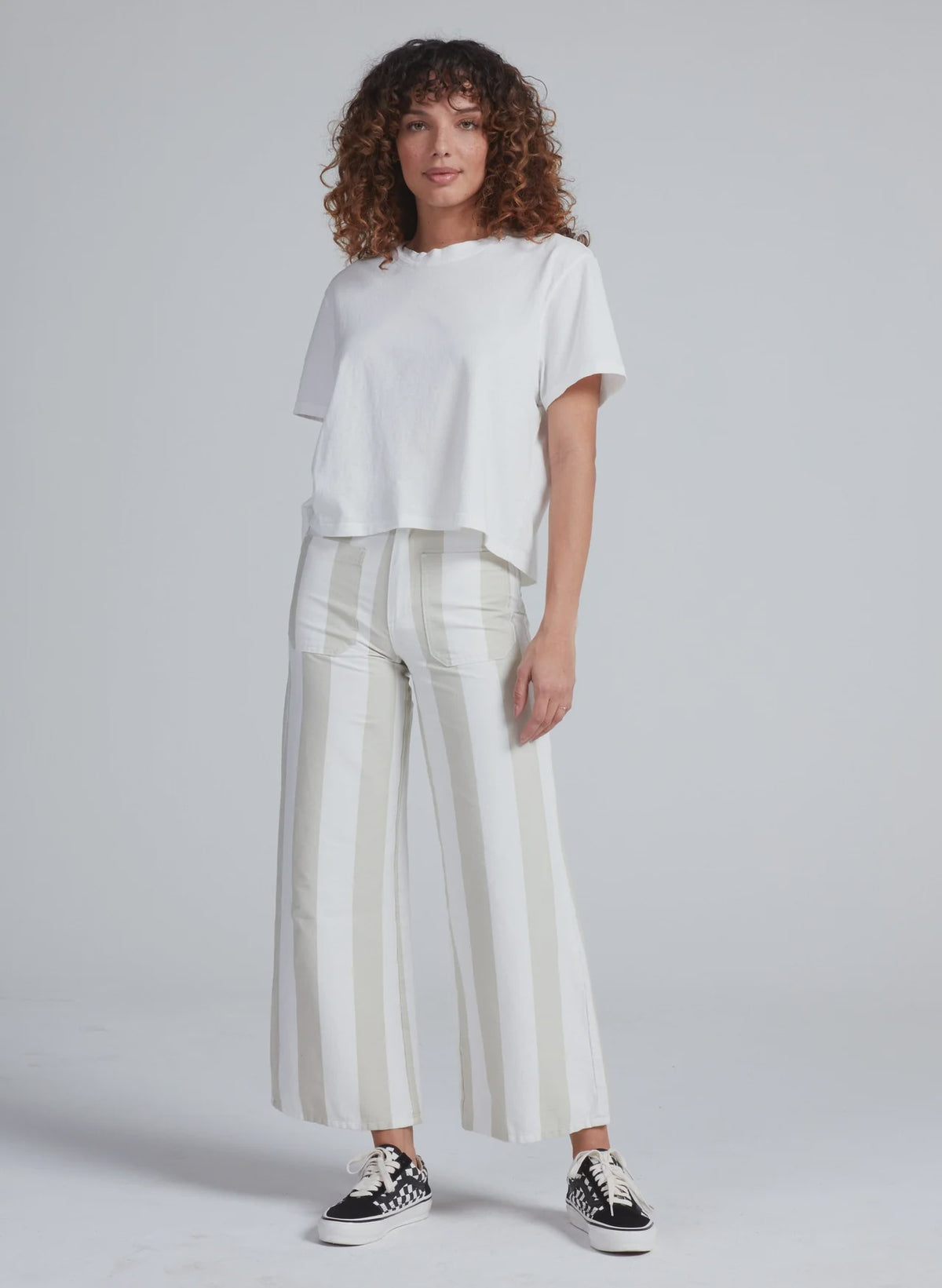 A person with curly hair wears a loose white T-shirt, ASKK’s Cropped Sailor high-rise wide-leg stretch twill pants with beige stripes, and black-and-white sneakers, standing against a plain light gray background.