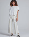 A person with curly hair wears a loose white T-shirt, ASKK’s Cropped Sailor high-rise wide-leg stretch twill pants with beige stripes, and black-and-white sneakers, standing against a plain light gray background.