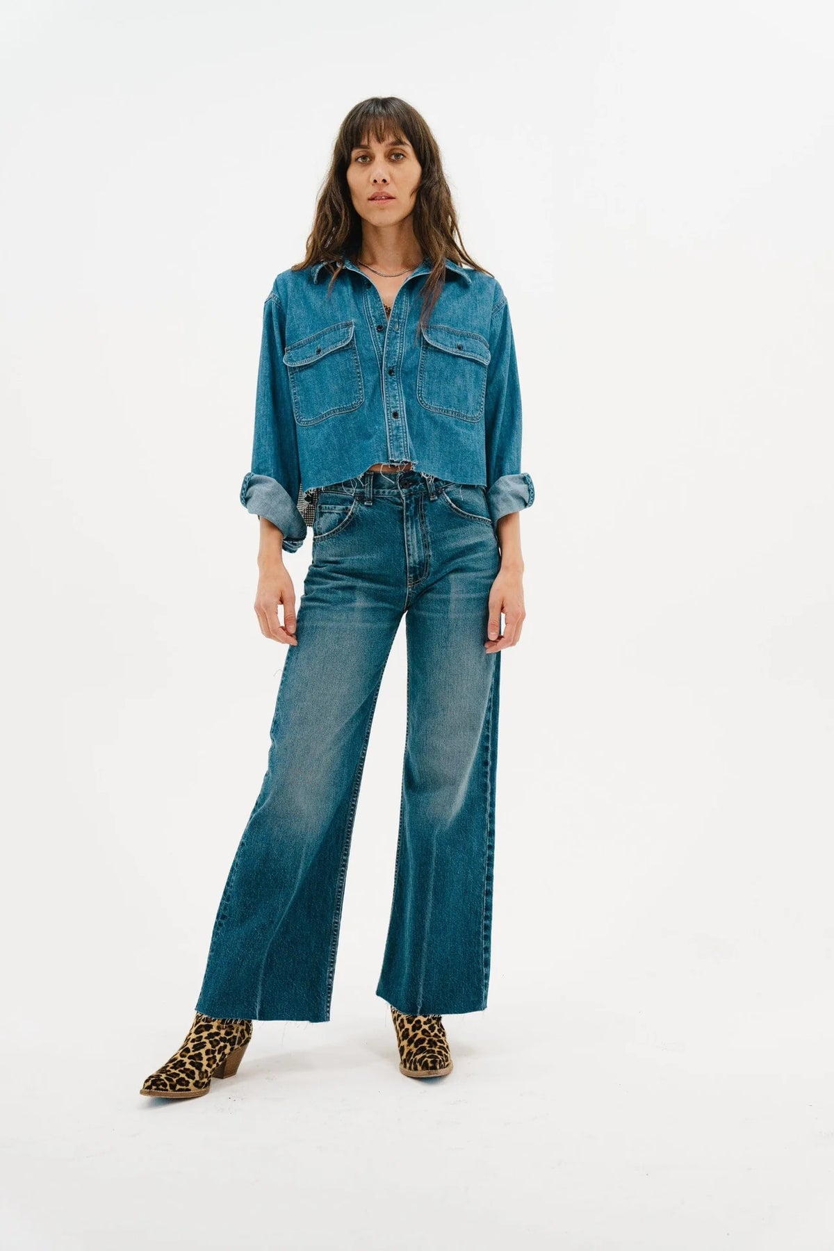 A woman poses against a white background in the ASKK Crosby Flannel, paired with rolled-up sleeves, high-waisted wide-leg jeans, and leopard print ankle boots. Her long brown hair frames her face, completing her chic look.