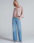 A person stands against a plain background wearing the ASKK Crosby Flannel—a cropped, long-sleeve plaid shirt—paired with light blue wide-leg jeans and checkered slip-on shoes. One hand is in their pocket, and they look relaxed.