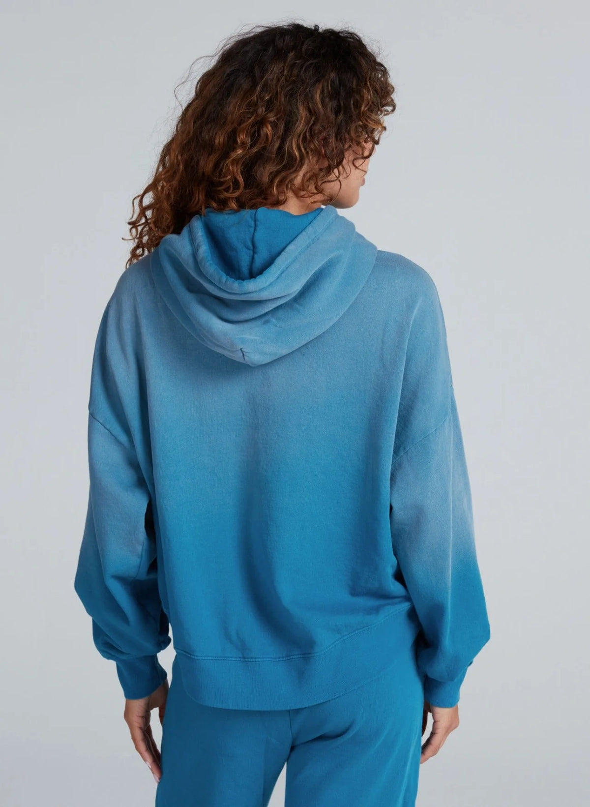 Person with curly hair shown from the back in ASKK's Indigo Dip Dye Hoodie and matching pants, both in a relaxed fit and made from soft American knit cotton, against a plain light background.