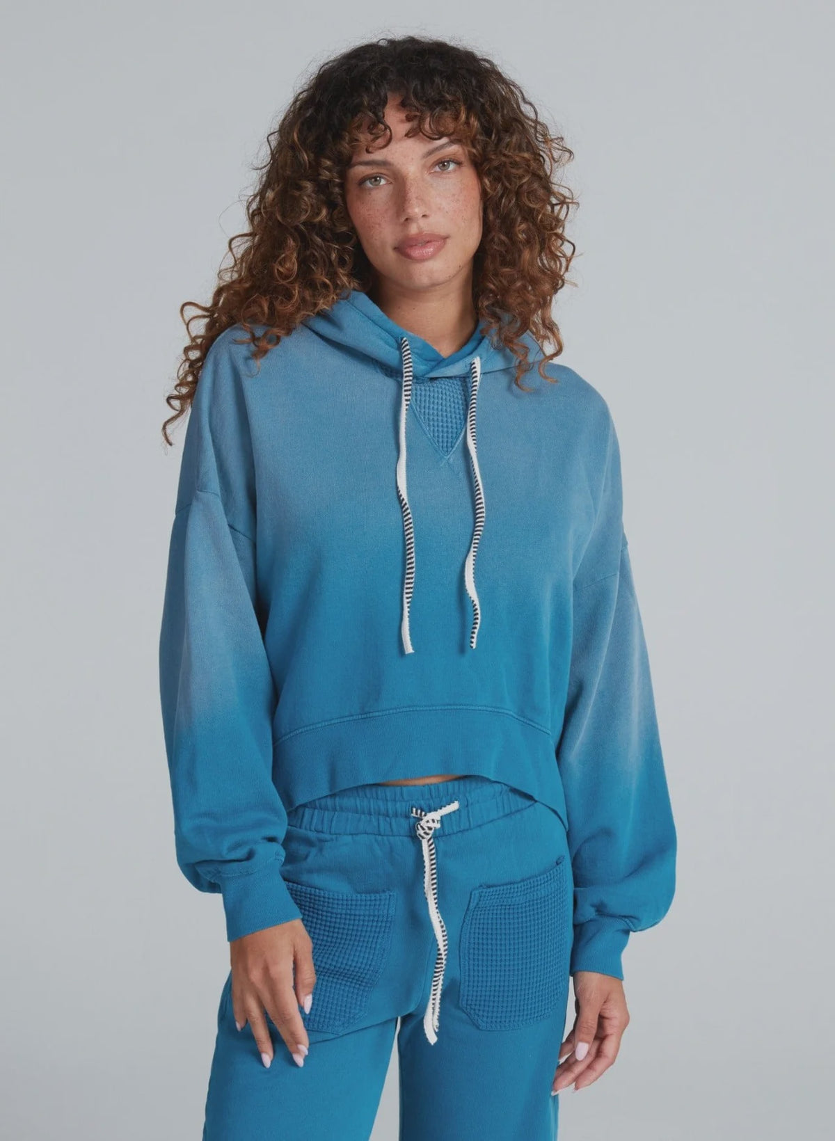 A woman with curly brown hair wears the ASKK Indigo Dip Dye Hoodie and matching joggers, featuring textured pockets and crafted from soft American knit cotton, standing against a plain light gray background.