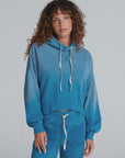 A woman with curly brown hair wears the ASKK Indigo Dip Dye Hoodie and matching joggers, featuring textured pockets and crafted from soft American knit cotton, standing against a plain light gray background.