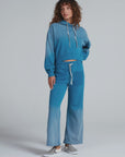 A person with curly hair wears ASKK Indigo Dip Dye Sailor Sweats, a blue gradient American knit cotton hoodie and matching wide-leg sweatpants, paired with black-and-white checkered slip-ons, against a light gray background. Made in Los Angeles.