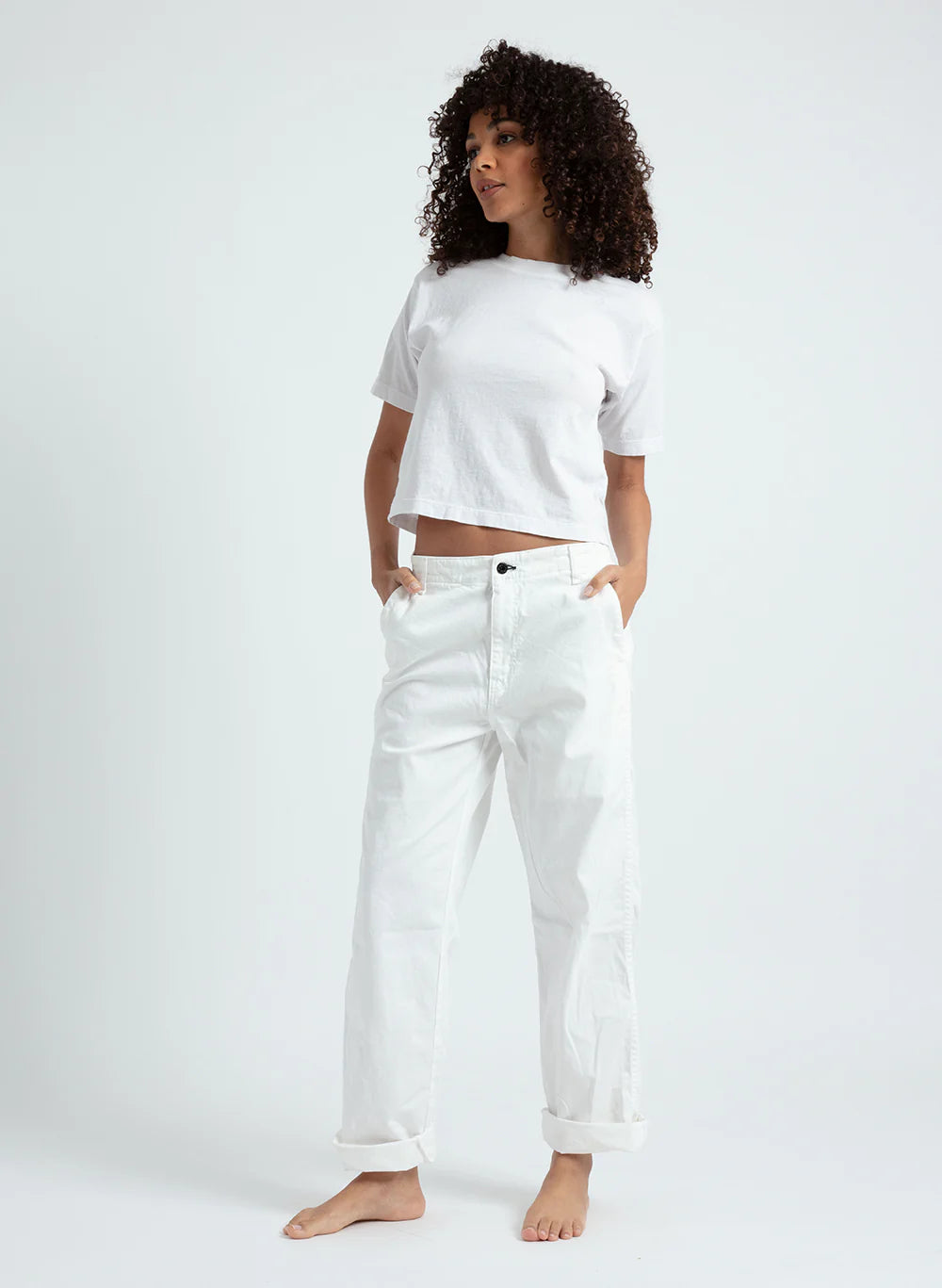 A woman with curly hair wears a white t-shirt and ASKK chino twill relaxed, loose fit pants, standing barefoot against a plain light background with her hands in her pockets, looking slightly to the side.