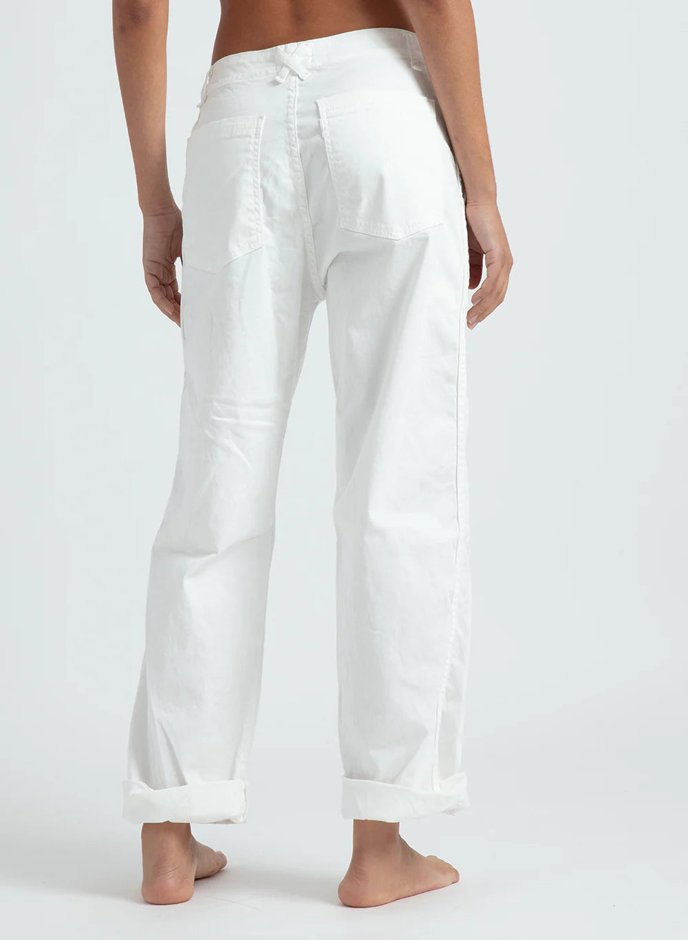 A person stands barefoot on a light-colored floor, facing away from the camera, wearing ASKK's relaxed-fit chino twill pants with rolled cuffs and back pockets for comfort.