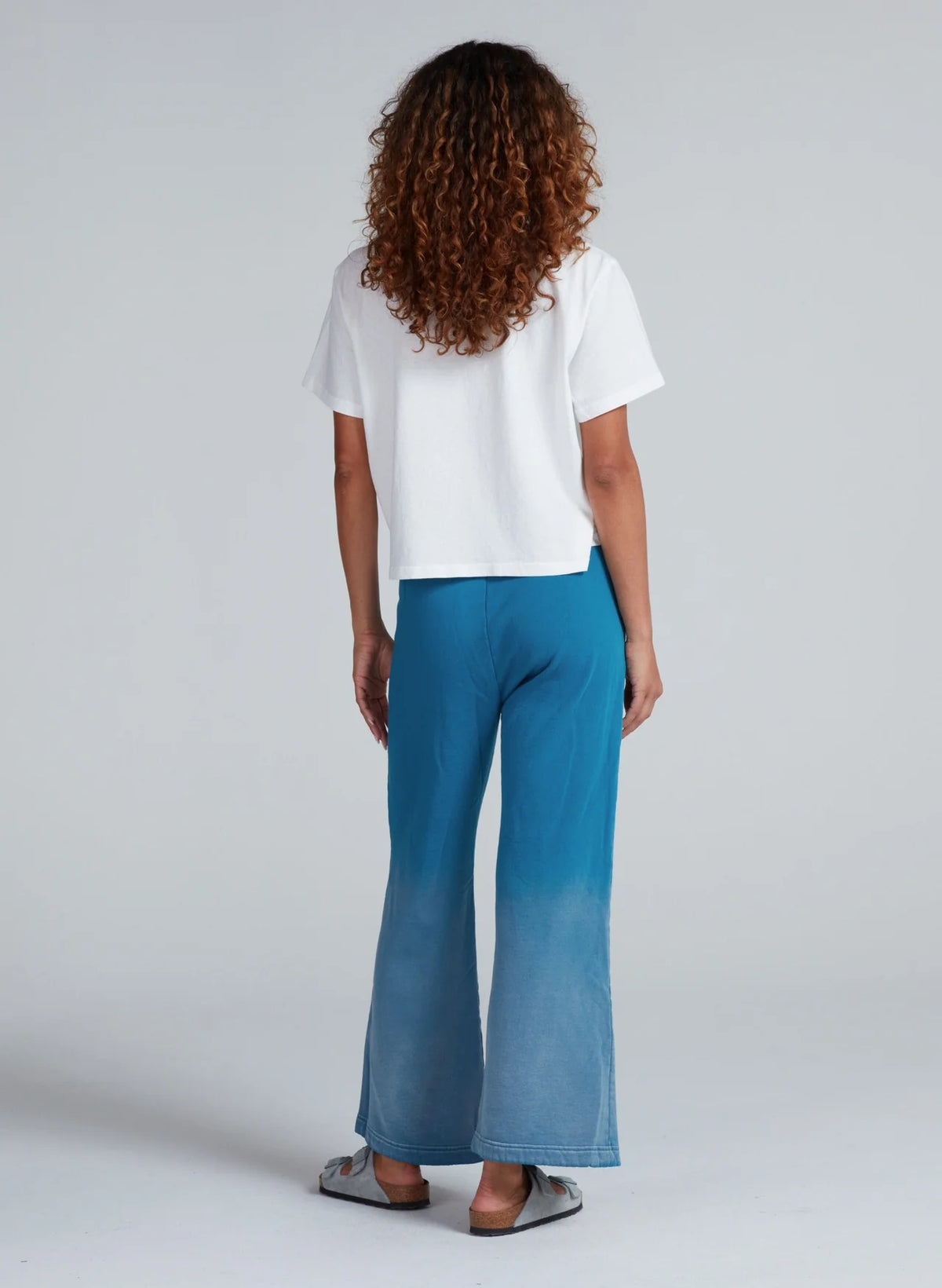 A person with curly hair stands facing away in a loose white short-sleeve top and ASKK Indigo Dip Dye Sailor Sweats, made from American knit cotton in Los Angeles, paired with gray slip-ons. The background is plain light gray.