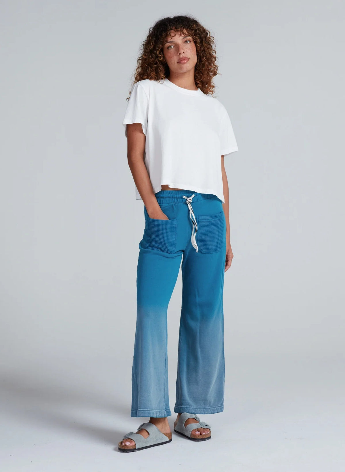 A person with curly hair wears a loose white t-shirt and ASKK Indigo Dip Dye Sailor Sweats, paired with gray sandals, standing against a plain light gray background.