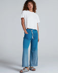 A person with curly hair wears a loose white t-shirt and ASKK Indigo Dip Dye Sailor Sweats, paired with gray sandals, standing against a plain light gray background.