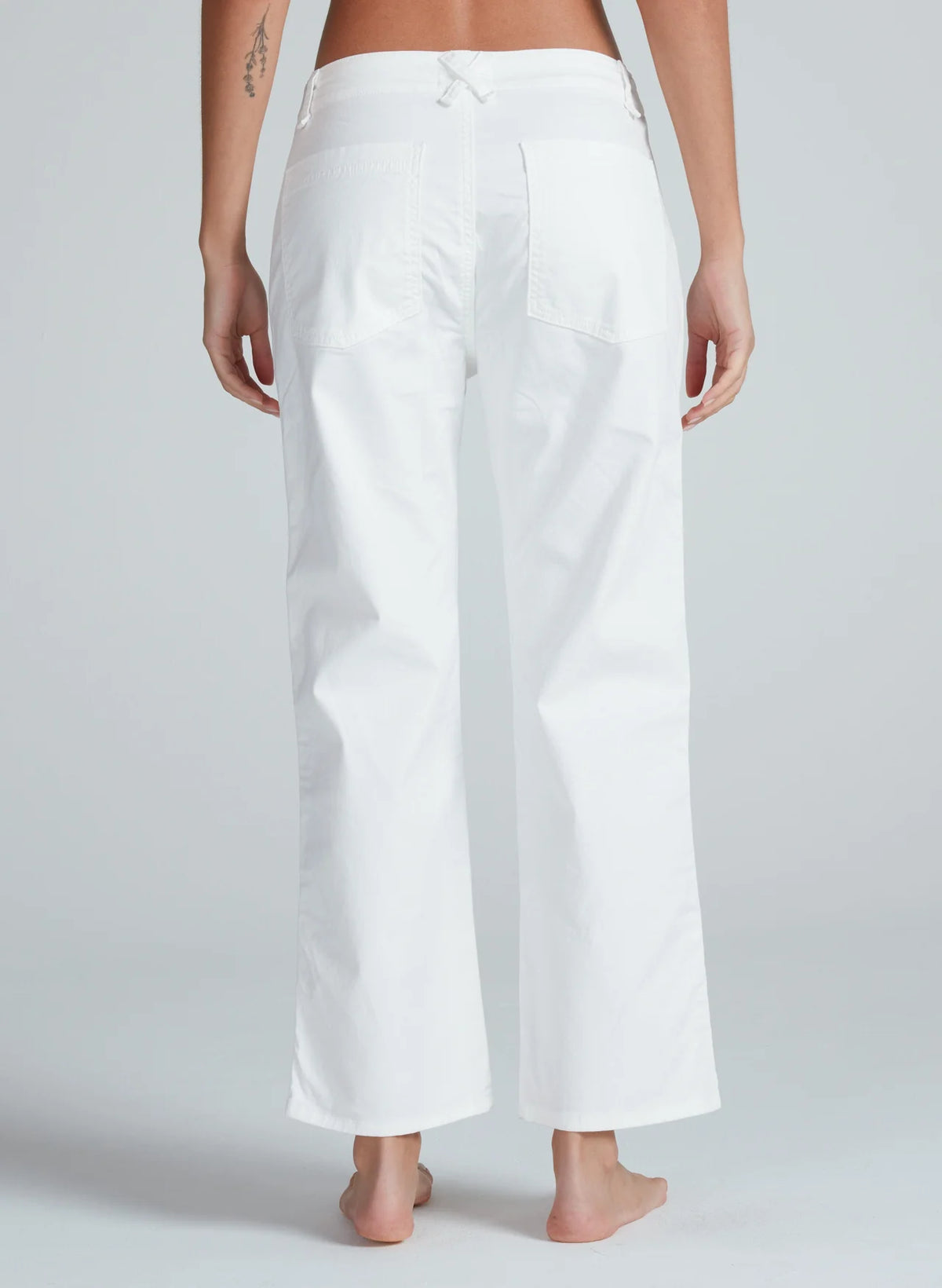 A person stands barefoot, facing away from the camera, wearing ASKK’s Tomboy Twill high-waisted, loose-fit white stretch twill pants with large back pockets and a straight leg against a plain light gray background.