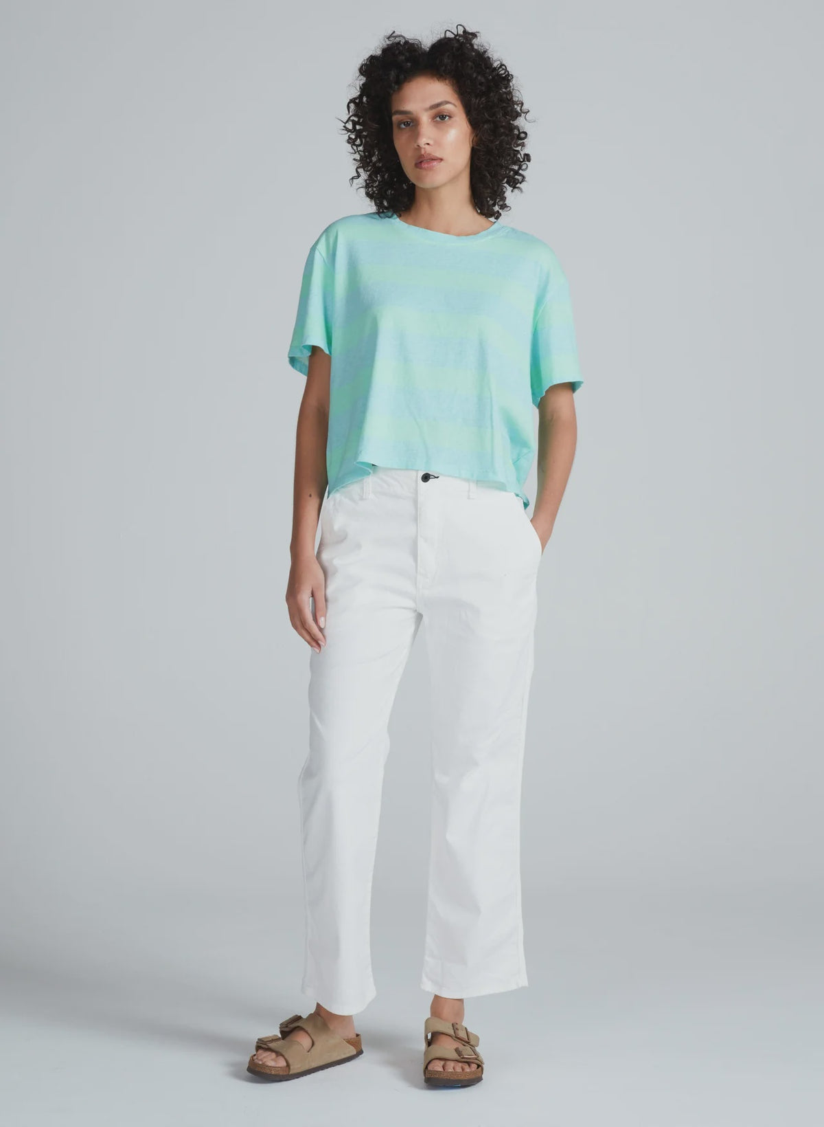 A person with curly hair wears a loose light blue-green t-shirt, ASKK's Tomboy Twill high waist white stretch pants, and beige sandals, standing against a plain light gray background.