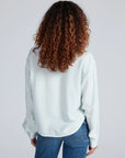 A person with curly brown hair is shown from the back wearing the ASKK Crosby Shirt—a relaxed-fit, light pinstriped long-sleeve shirt—with blue jeans, set against a plain light background.