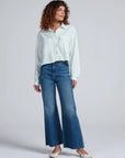 Wearing the ASKK Crosby Shirt, a woman with curly hair pairs it with blue high-waisted wide-leg jeans and cream flats, standing against a plain light background.
