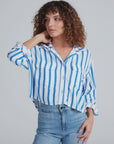 A person with curly brown hair wears the ASKK Crosby Shirt—blue and white vertical stripes, 100% cotton, relaxed fit—and light blue jeans, posing with one hand touching their neck against a plain light background.
