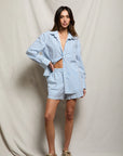 A woman poses against a neutral draped backdrop in Perfectwhitetee's Julie Poplin Short and matching striped button-up shirt, paired with beige sneakers. One hand on her hip, the partly unbuttoned shirt completes this effortless summer look.