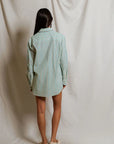A person with long dark hair, wearing the Perfectwhitetee Avalon Poplin Button Down in blue and white stripes and white slip-on shoes, stands with their back to the camera against a light draped backdrop.