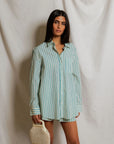 A woman stands before a white draped backdrop, wearing the Avalon Poplin Button Down by Perfectwhitetee with matching shorts. She holds a beaded handbag in her left hand and gazes toward the camera.