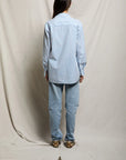 A person with long dark hair faces away, wearing the Avalon Poplin Button Down by Perfectwhitetee in blue and white stripes, paired with light blue jeans and beige shoes. Set against draped light fabric—an ideal chic summer cover-up look.