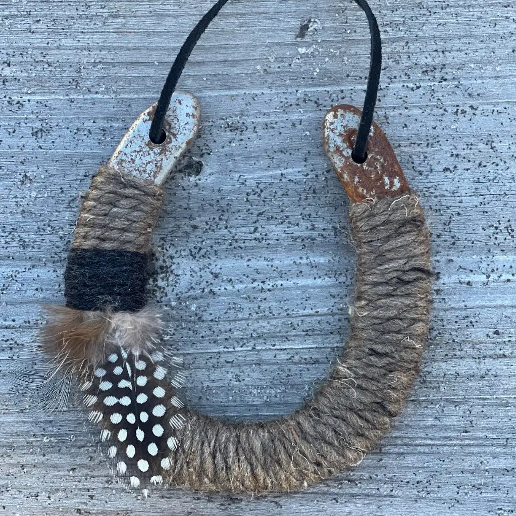 The Lucky Horseshoe by Faire features an upcycled horseshoe wrapped in twine with a black band and spotted feather, hanging from a black cord—a unique housewarming gift displayed against rustic wood.