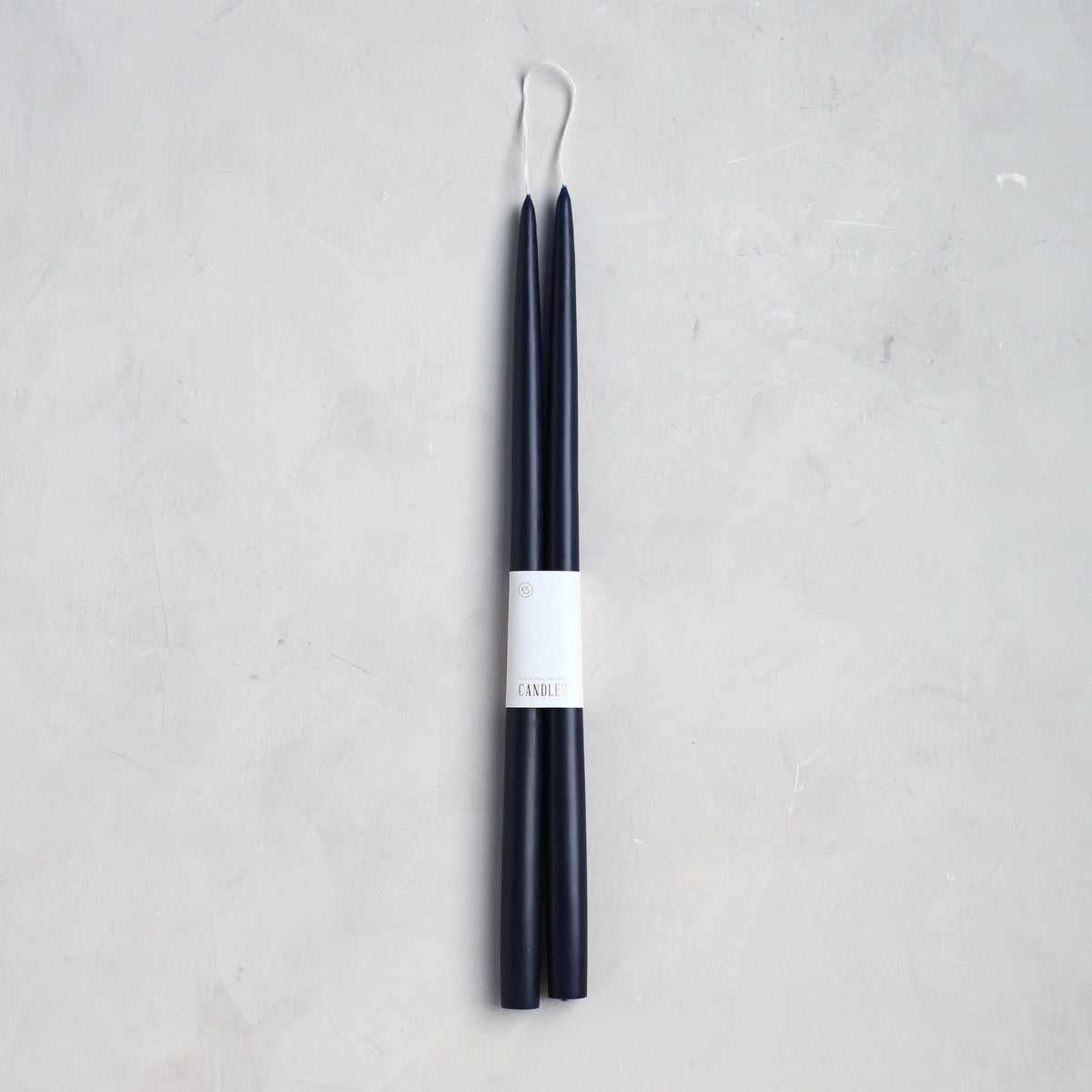 The Floral Society’s 18" Taper Candle - Pair features two tall, slender dark blue candles joined by a wick and wrapped with a white “CANDLE” label, ideal for elegant home décor. Displayed on a light gray textured surface.
