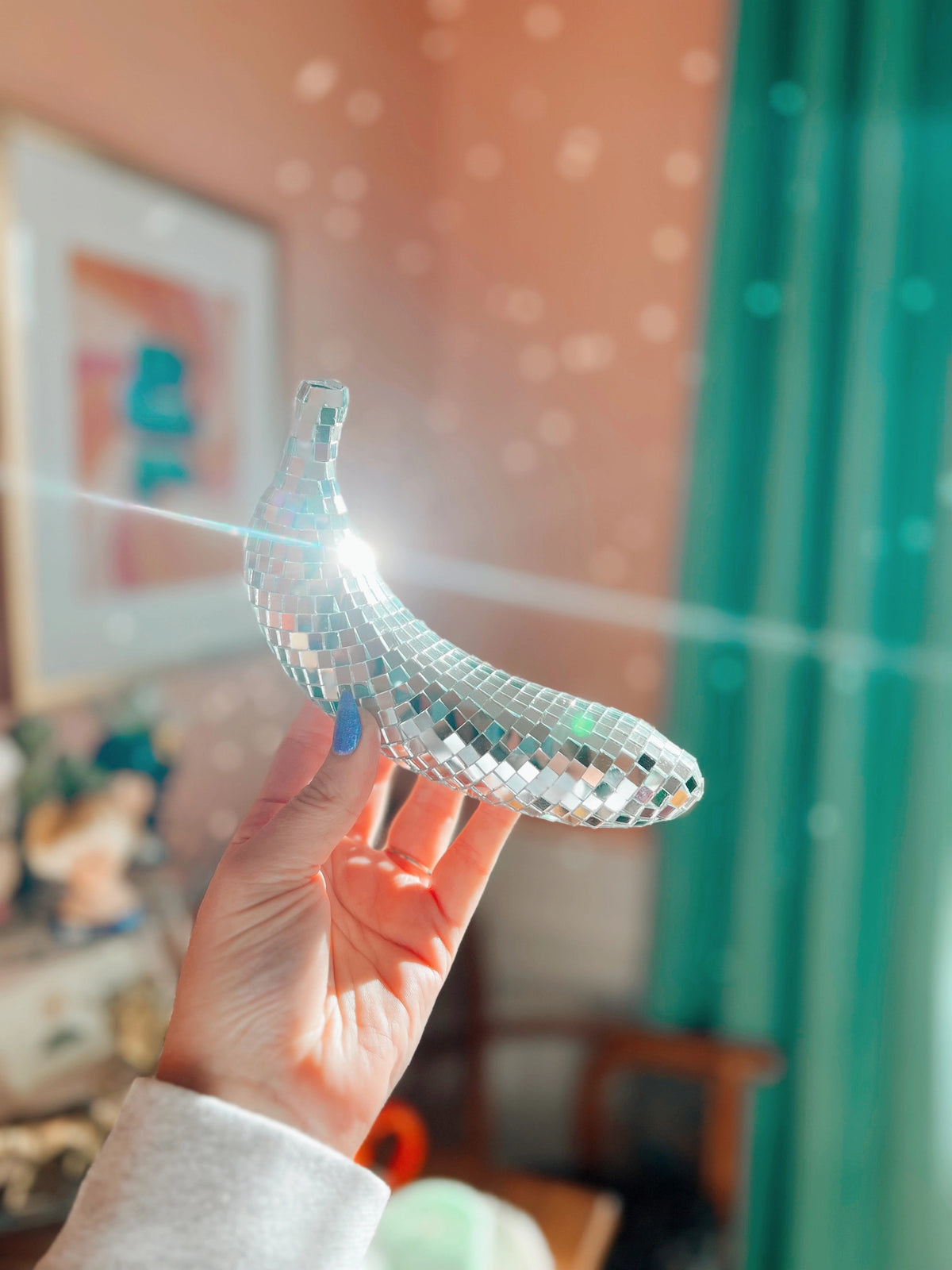 A hand holds up the Faire Disco Banana, a handmade piece covered in tiny mirror tiles that reflect sunlight around a cozy, colorful room with green curtains and framed art.