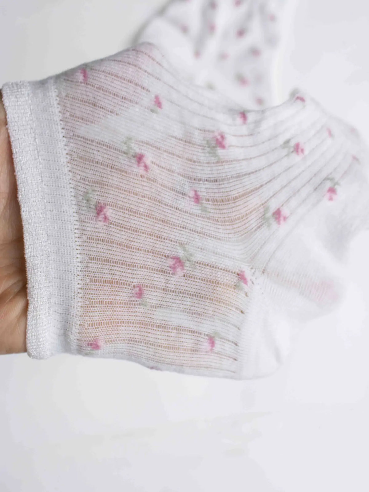 A hand holds up Faire's Blossom Breeze Sheer Floral Ankle Socks, featuring thin, ribbed, slightly transparent fabric with delicate pink and green floral patterns—ideal for those wanting lightweight, breathable socks with a feminine touch.