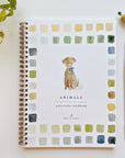 Faire's "Watercolor Workbook," titled "Animals," rests on a surface. It features beginner-friendly illustrations and a spiral binding for easy page turns. The cover, with vibrant squares, displays a painted dog with a blue bow. Nearby lie a paintbrush, paint tube, and green-leafed plant.