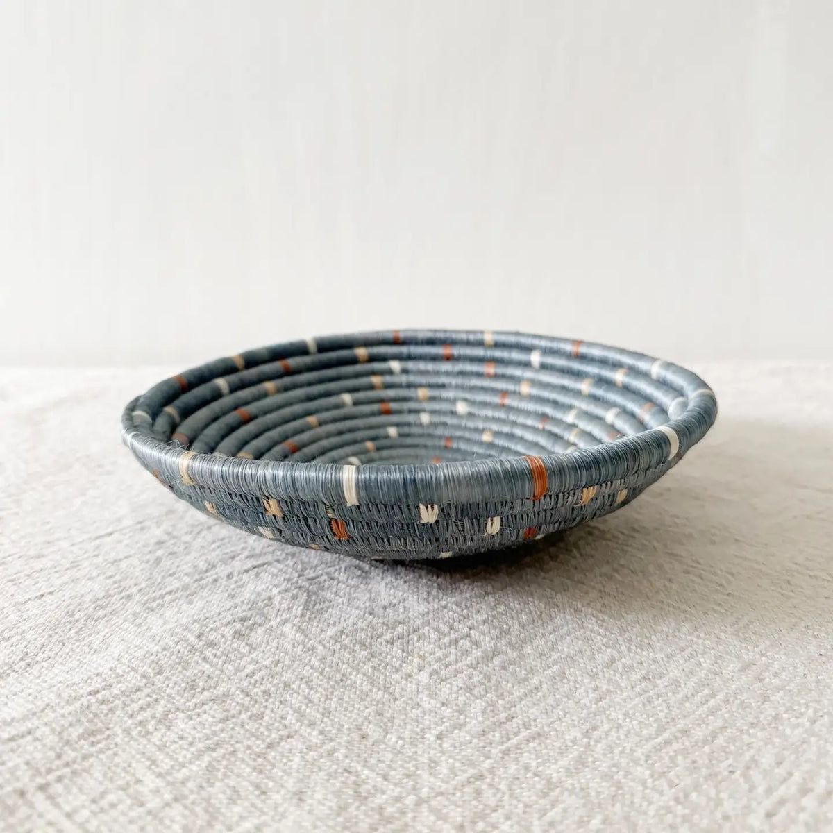 The Kitopo Small Bowl by Faire, a shallow, round Rwandan basket with blue and gray coils plus white and orange accents, rests on textured light gray fabric against a neutral background.