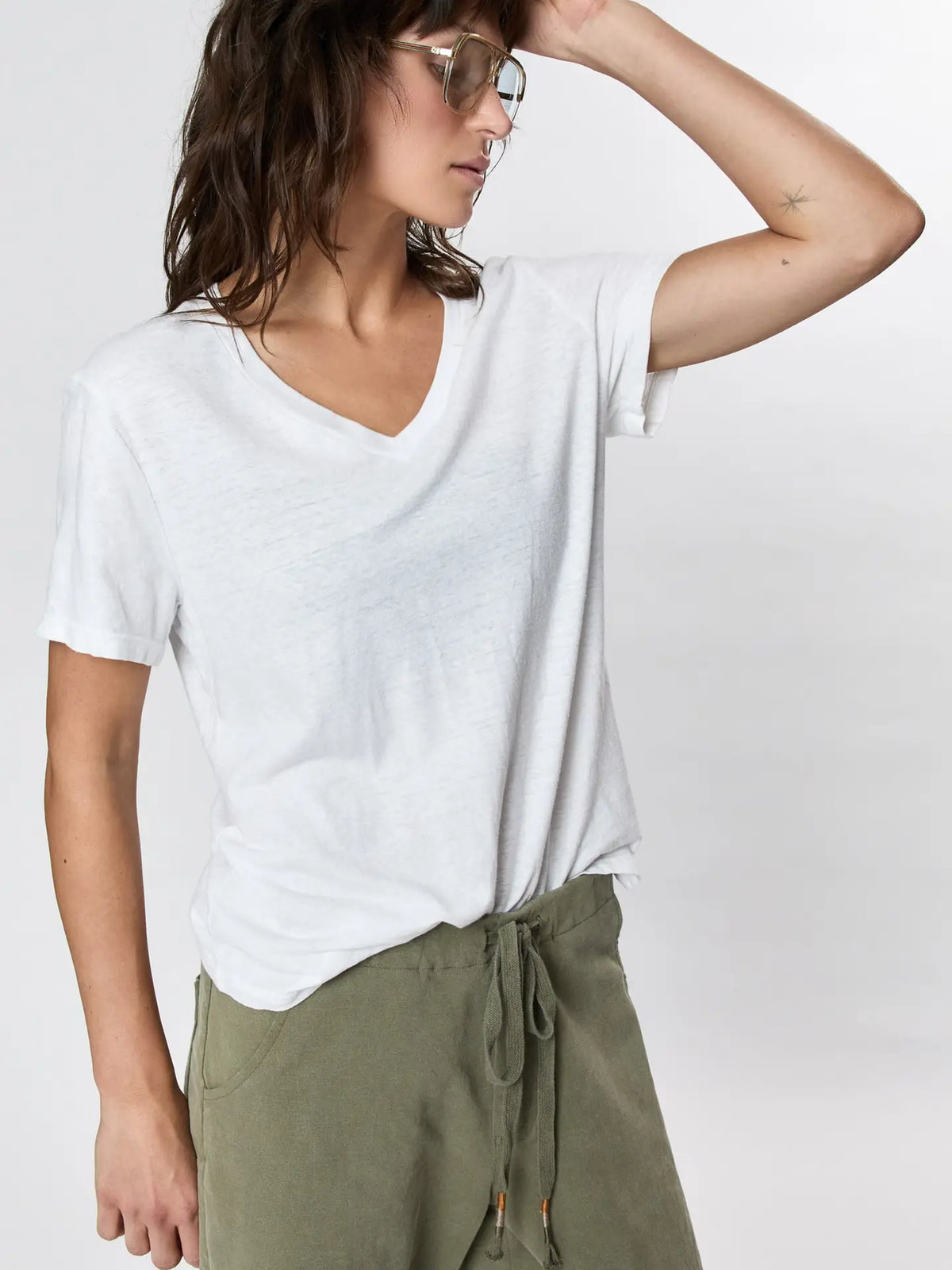 Wearing Faire’s Hemp V Neck in white—a loose, vintage-fit T-shirt—paired with olive green drawstring pants and sunglasses, a person stands against a plain light background, one hand resting on their forehead.