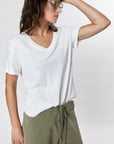 Wearing Faire’s Hemp V Neck in white—a loose, vintage-fit T-shirt—paired with olive green drawstring pants and sunglasses, a person stands against a plain light background, one hand resting on their forehead.