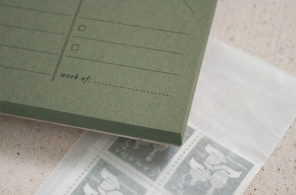 A green Weekly Planner Paper Notepad by Faire, labeled "week of:", sits partially atop a white envelope filled with vintage gray postage stamps.