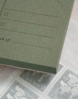 A green Weekly Planner Paper Notepad by Faire, labeled "week of:", sits partially atop a white envelope filled with vintage gray postage stamps.