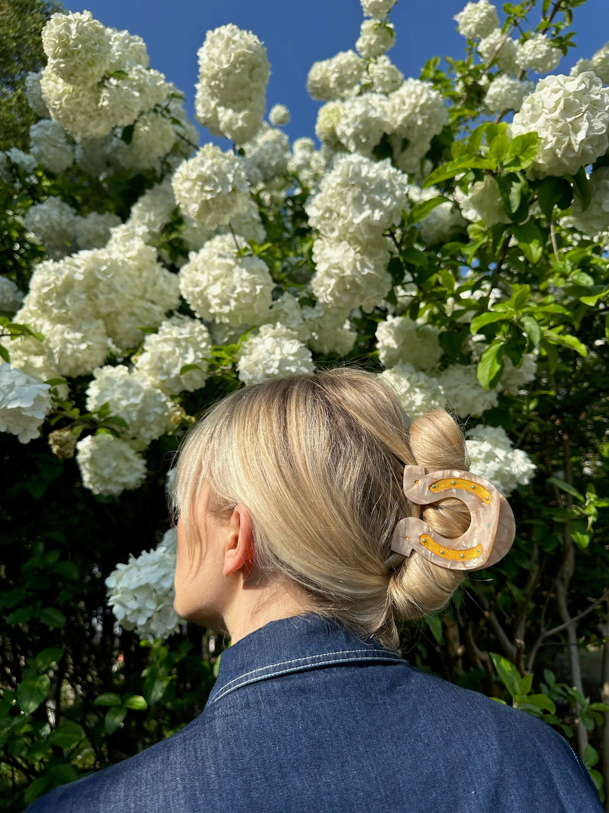 Faire Lucky Horseshoe Hair Clip on blonde hair, styled outdoors among blooming white flowers and green foliage.