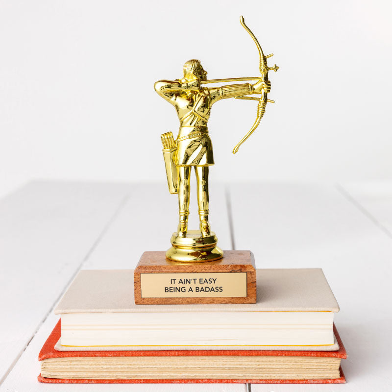 A golden JE Trophy depicting a figure with a bow and arrow, standing on a stack of books in Arizona style. The base of the trophy has a plaque that reads "IT AIN'T EASY BEING A BADASS." (Faire)