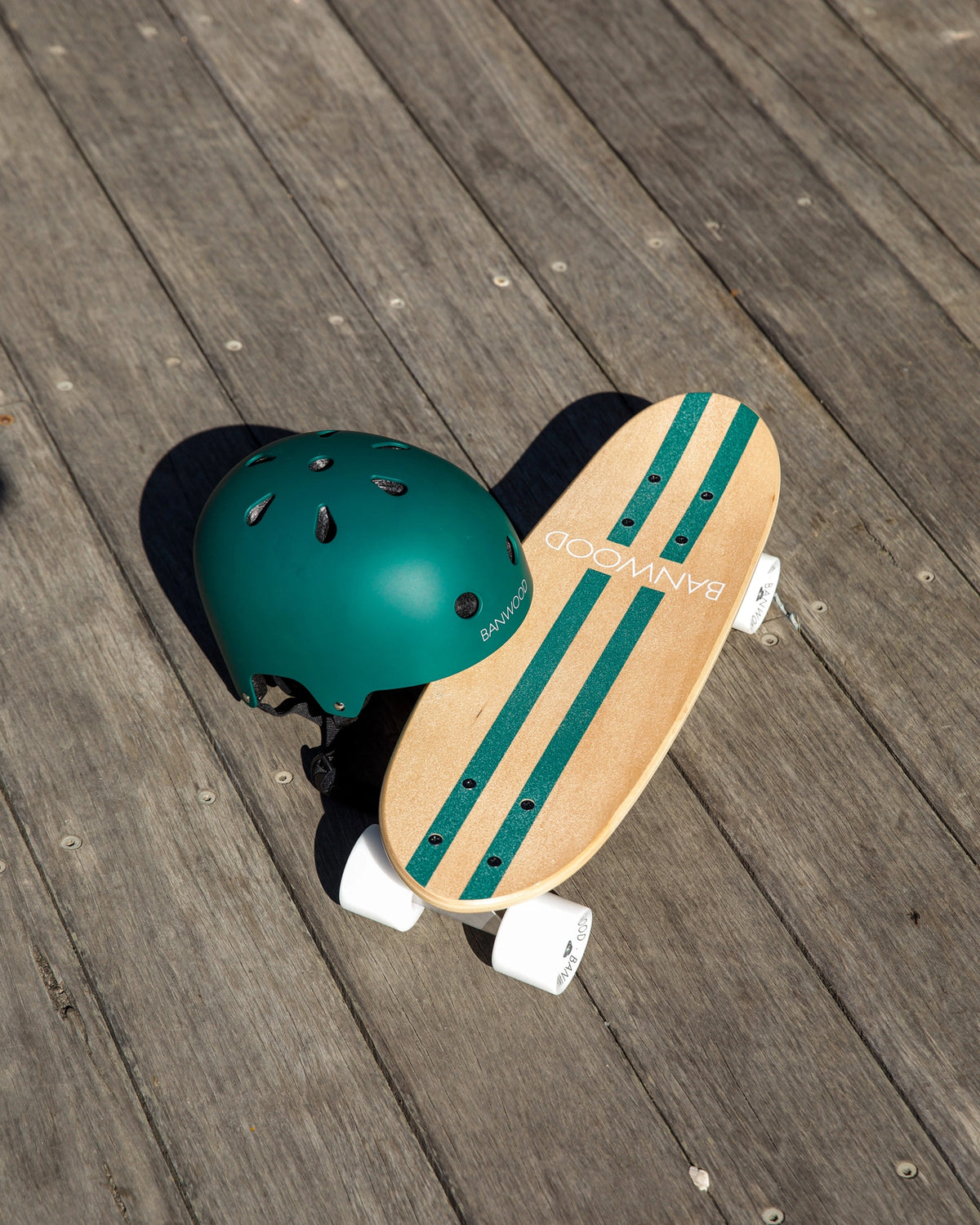 A green helmet and a BANWOOD SKATEBOARD - GREEN by Faire rest on a wooden deck in sunlight. The board features white wheels, Canadian maple construction, and "BAMBOO" printed on its surface.