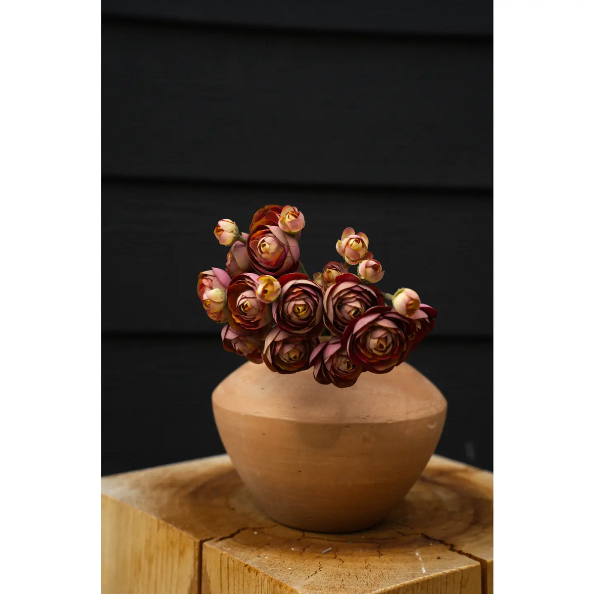 The Faire Ranunculus Bouquet features dark pink and cream faux ranunculus stems beautifully arranged in a round brown clay vase, displayed on wood with a black background.