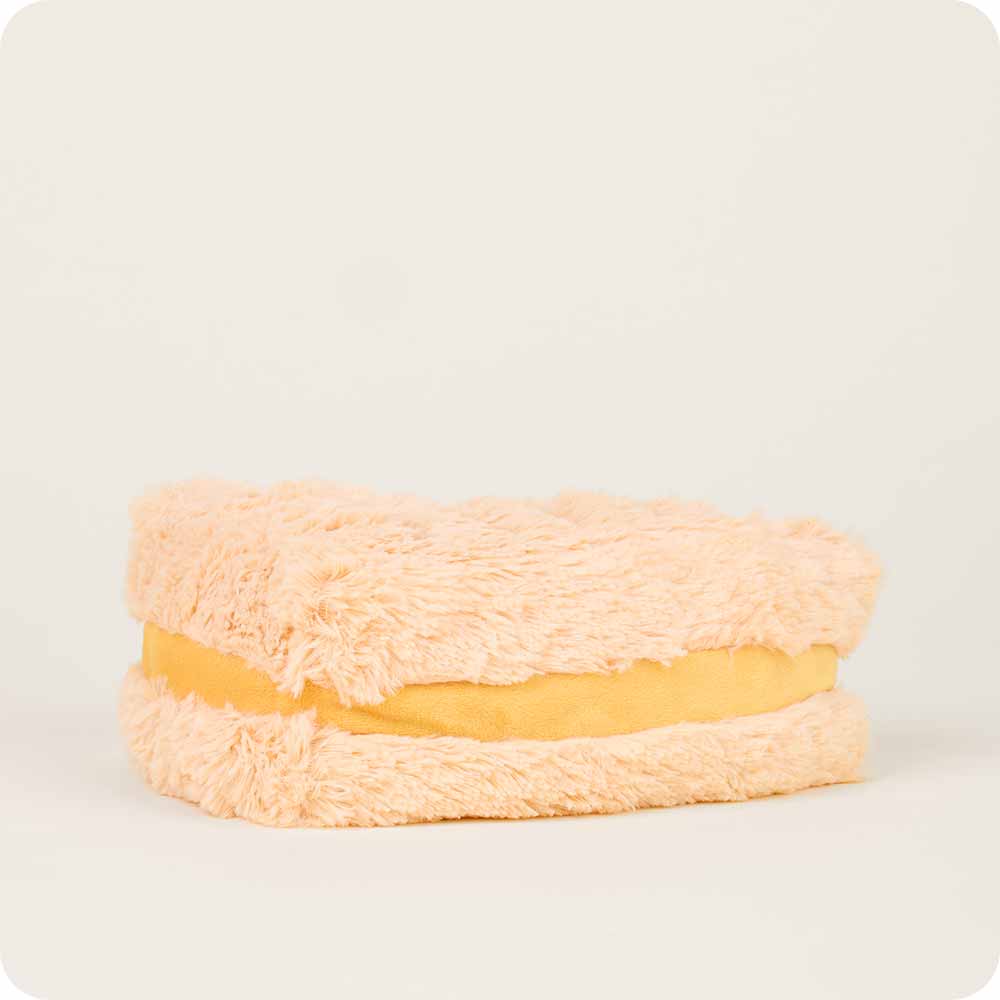 The Grilled Cheese Warmies by Faire is a plush, beige sandwich-shaped pillow with a fluffy texture and yellow center, designed to be microwavable for cozy warmth—perfectly resembling a classic grilled cheese.
