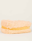 The Grilled Cheese Warmies by Faire is a plush, beige sandwich-shaped pillow with a fluffy texture and yellow center, designed to be microwavable for cozy warmth—perfectly resembling a classic grilled cheese.