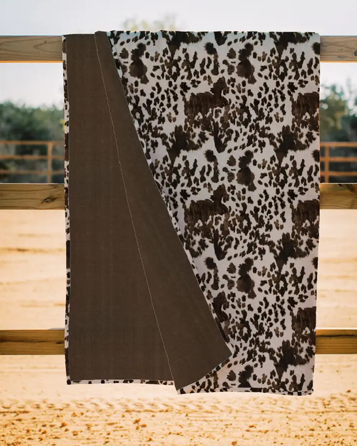 The Faire Bella Hide Brown Quilt, featuring a brown and white cowhide pattern with a solid brown underside, hangs over a wooden fence outdoors, bringing Fort Worth Texas style to any setting.