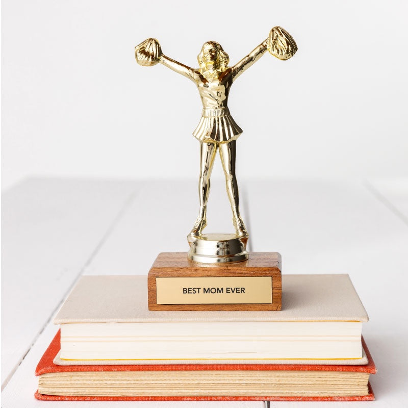 A JE Trophy depicting a woman with arms raised high, standing on a wooden base labeled "BEST MOM EVER", placed atop a stack of books in Bungalow style on a white background. (Brand: Faire)