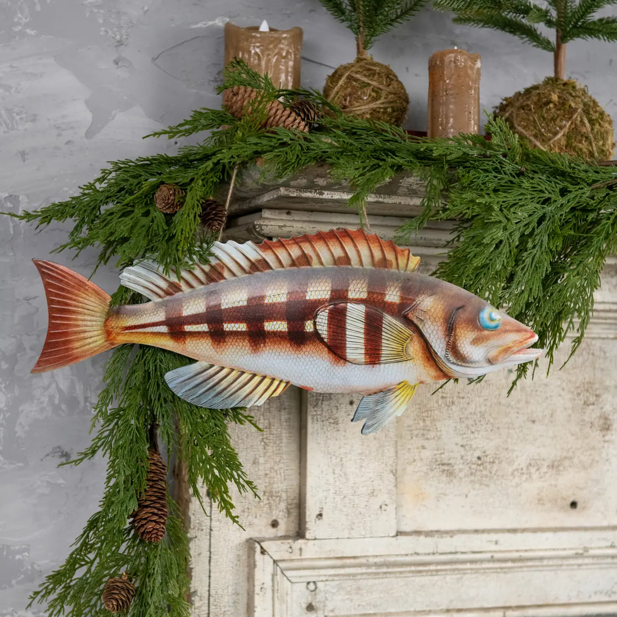 Walleye fish wall decor by The Faire hangs above a white mantel with greenery and candles for a cozy accent.
