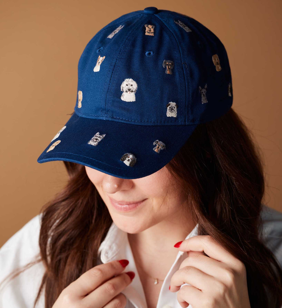 A person with long brown hair smiles slightly, wearing the Faire Dog Days Baseball Cap—a blue cap embroidered with small dog faces and an adjustable strap. The plain background is light brown.