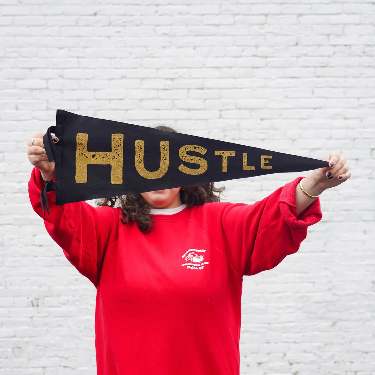 A person in a red sweatshirt holds the Faire Hustle Pennant, showcasing bold gold "HUSTLE" lettering and concealing their face. The backdrop is decorated with white brick wall decor.