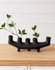 The Faire Black Crescent Shape ceramic vase with five openings, filled with green leafy stems, sits on a wooden surface against a white shiplap wall, creating striking decor.