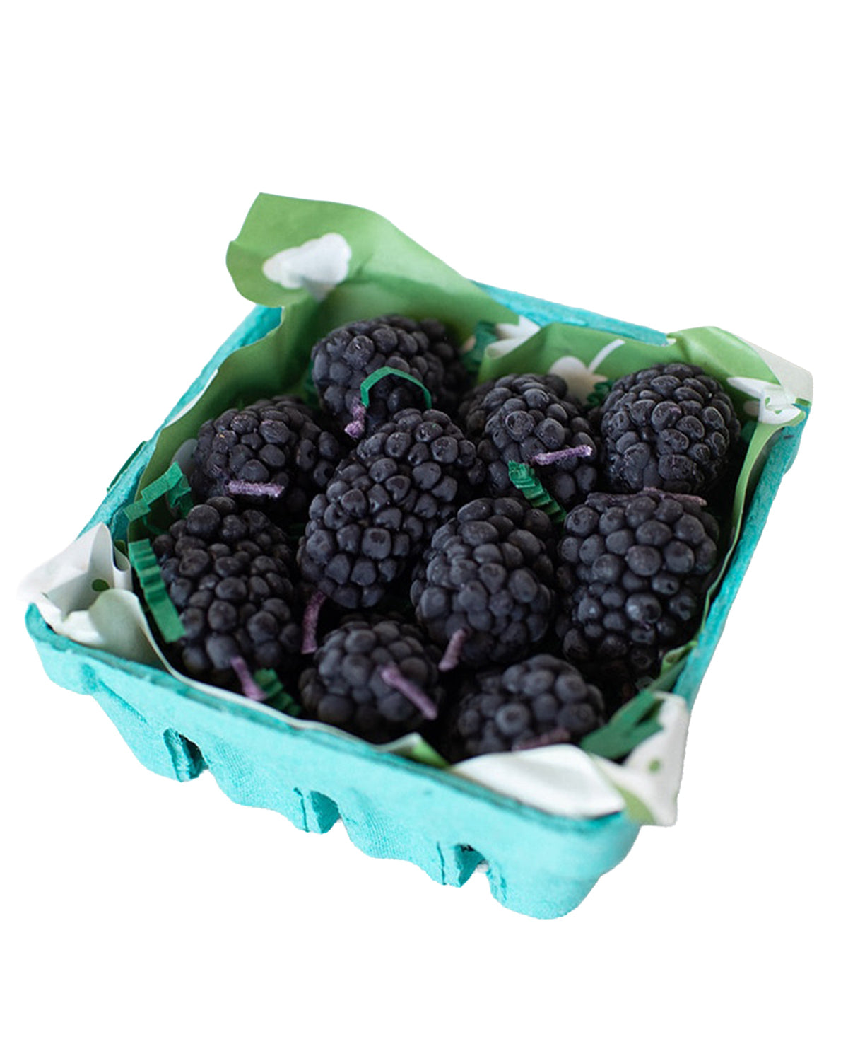 A small turquoise carton of fresh blackberries sits beside Faire’s Blackberry Beeswax Candles, both lined with green and white paper and set against a white background.