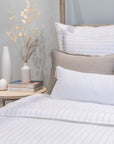 A cozy bedroom with a neatly made bed featuring sophisticated stripe bedding from the Lake Linen Collection and a Blake Big Pillow 28x36 by POM POM. Two stacked books and a white vase with dried flowers sit on a round wooden side table next to the bed, creating a setting with calm and minimalist decor.