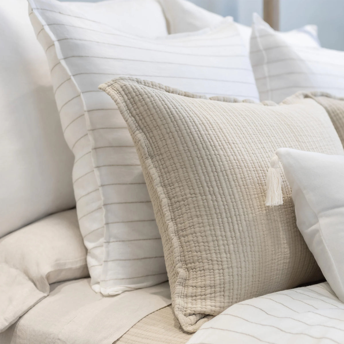 A neatly arranged bed adorned with a mix of textured pillows from POM POM's Lake Linen Collection, highlighted by the Blake Big Pillow 28x36 in beige with quilted tassels in the foreground. Smooth, white pillows featuring a sophisticated stripe pattern are positioned in the background, adding to the cozy and elegant atmosphere.