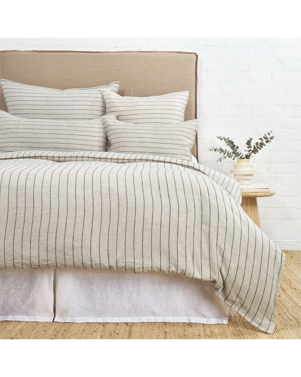 A neatly made bed showcases the POM POM Blake Duvet in beige and white stripes with matching pillows. A beige upholstered headboard and nightstand with a vase of greenery complete the relaxed look against a white brick wall.