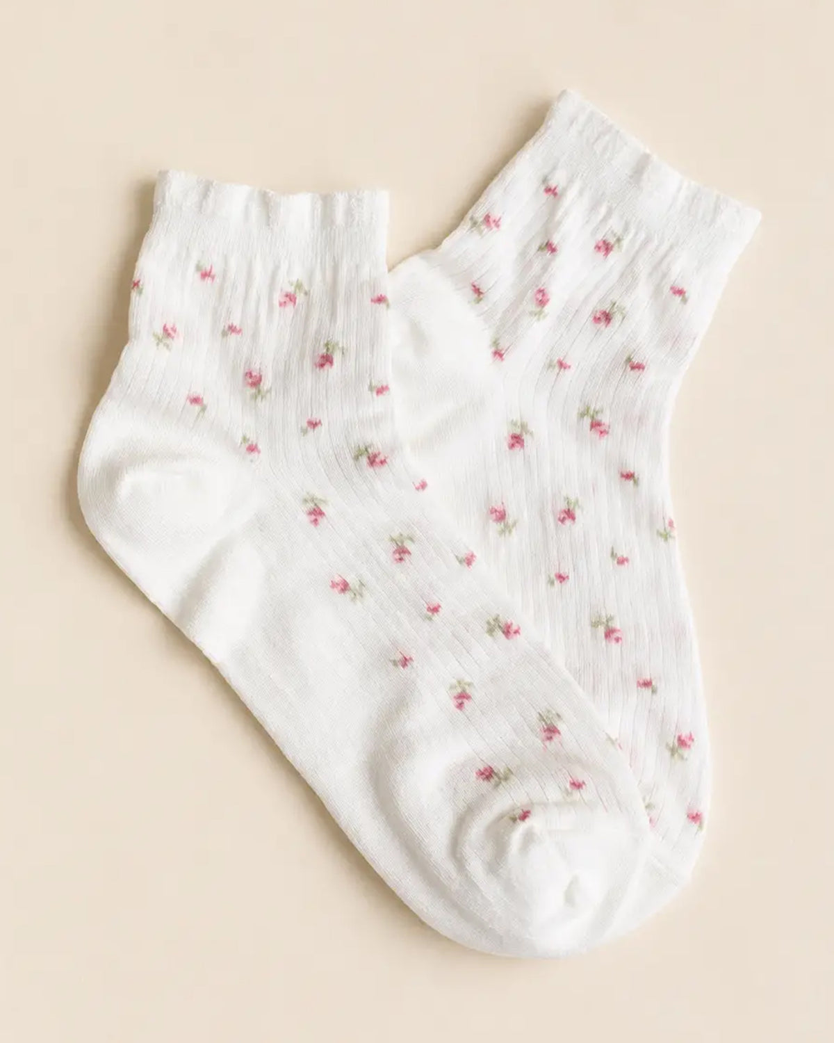 The Faire Blossom Breeze Sheer Floral Ankle Socks are white ankle socks with a subtle ribbed texture and small pink floral designs, shown laid flat on a light beige background.